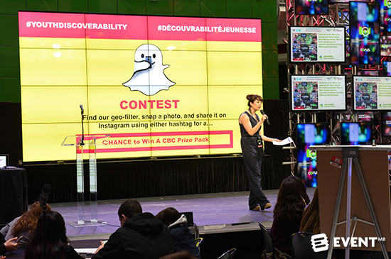 Person speaking with a screen behind her that says there is a contest for the first person to snapchat with a geofilter and hashtag on instagram or facebook gets a prize.